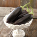 Three fresh, dark purple eggplants with green stems in a white decorative bowl on rustic wood. Healthy produce.