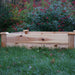 Cedar raised garden bed on green grass. Backyard gardening planter with vibrant red flowers in the background.