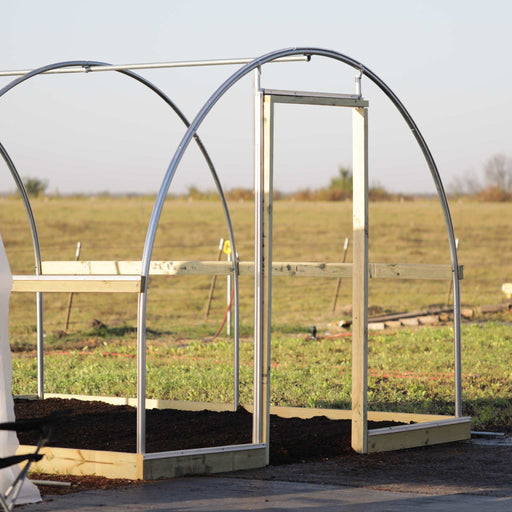 High tunnel greenhouse frame under construction with metal arches, wooden base, door frame, and raised garden beds.