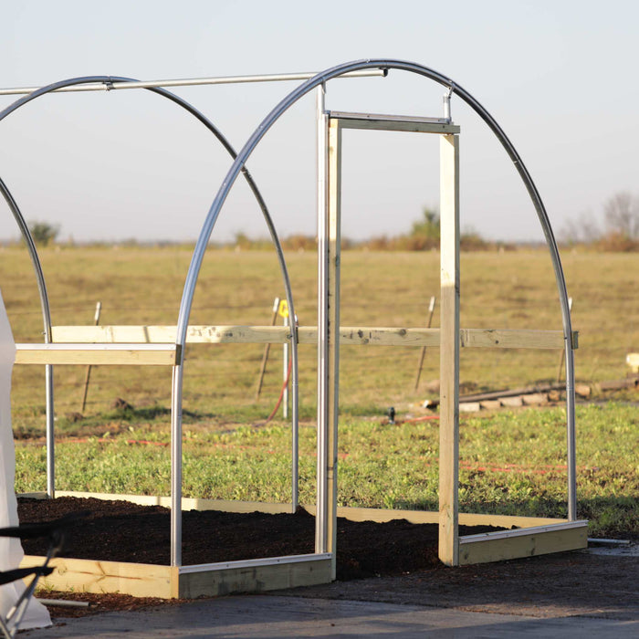 High tunnel greenhouse frame under construction with metal arches, wooden base, door frame, and raised garden beds.