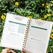 Garden planner with weekly habit tracker, goals, and pollinator observations, held in a vibrant flower garden.