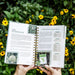 Hands hold a spiral-bound gardening planner showing muskmelon planting tips and warm season seed order list, with yellow flowers.