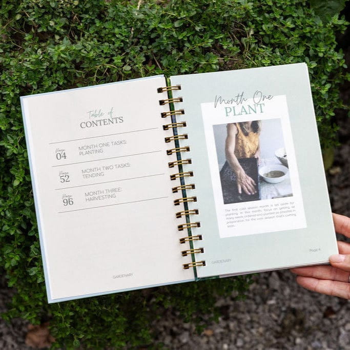 Gardenary spiral gardening planner open to "Table of Contents" (planting, tending, harvesting) & "Month One Plant" guide on foliage.