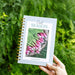 Cool Season Gardenary planner with radishes on cover, held in a hand, surrounded by lush green garden foliage.