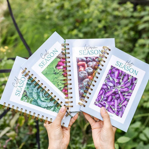 Hands holding four seasonal garden planners: Cold, Cool, Warm, Hot Season. Covers show broccoli, radishes, tomatoes, eggplants.
