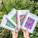 Hands holding four seasonal garden planners: Cold, Cool, Warm, Hot Season. Covers show broccoli, radishes, tomatoes, eggplants.