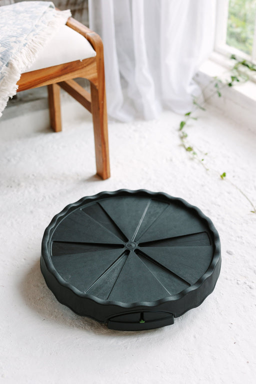 Dark gray GreenWay mini-trolley plant caddy on white floor. Wooden chair, patterned blanket, window with white curtains & greenery.