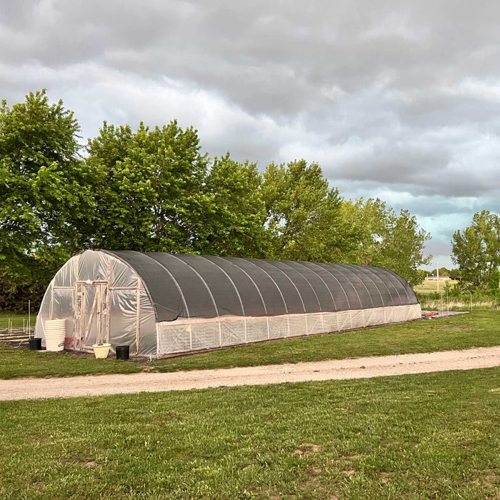 High tunnel greenhouse with black shade cloth roof, clear sides, and dirt path in a green field under cloudy sky.
