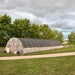 High tunnel greenhouse with black shade cloth roof, clear sides, and dirt path in a green field under cloudy sky.