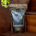 Azomite Natural Trace Minerals soil amendment, 2 lbs, on wood shelf with blurred yellow flowers in background.