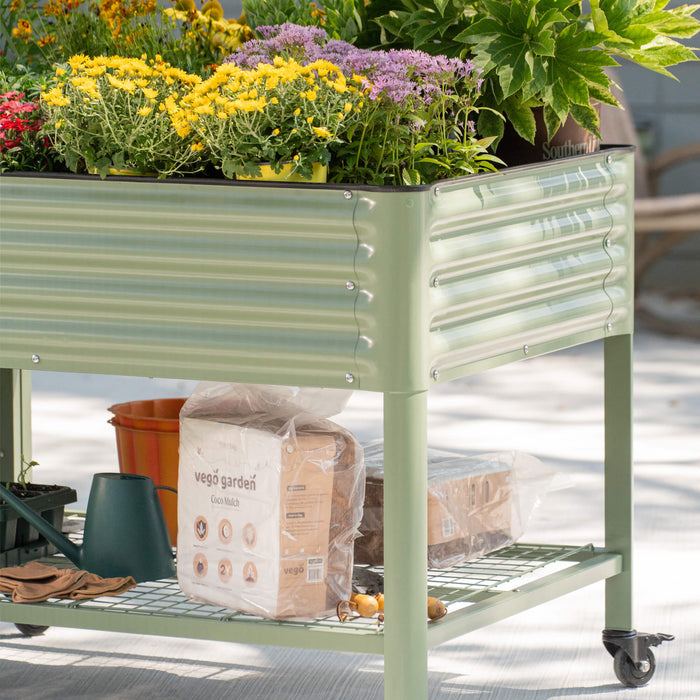 Sage green metal mobile raised garden bed filled with colorful flowers. Gardening tools and coco mulch on lower shelf.