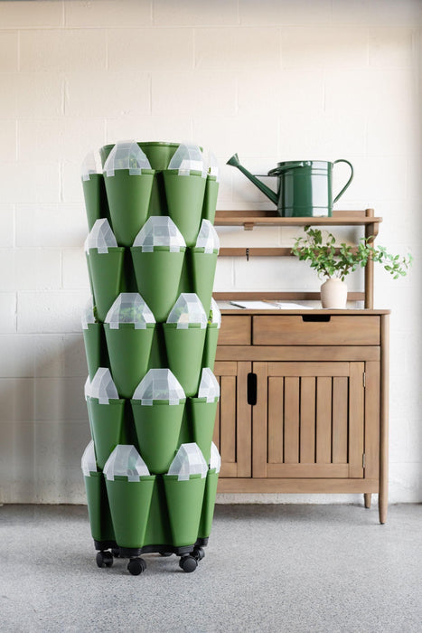 Green vertical garden tower on wheels with plants & clear covers. Wooden cabinet, watering can. Indoor urban gardening.