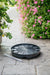 Black self-watering planter base, water level tube on stone patio. Strawberry plant, vibrant pink petunias in garden background.