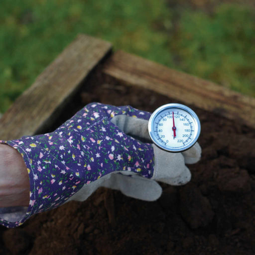 Gardener's gloved hand holding a compost thermometer, measuring temperature in a compost pile.