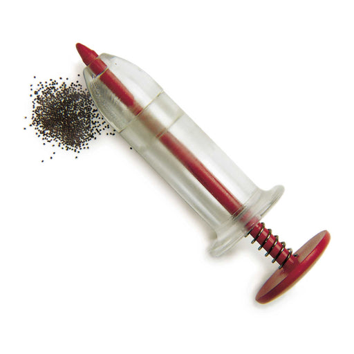 Garden seed sower, clear plastic with red spring plunger, beside small dark seeds for planting.