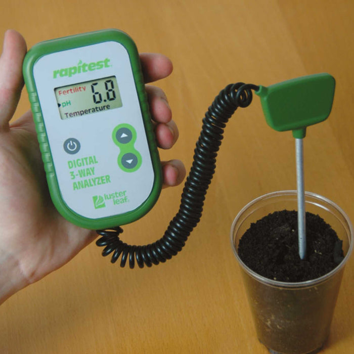 Rapitest digital soil pH, fertility, temperature analyzer (showing 6.8 pH) held, with probe inserted into dark soil.