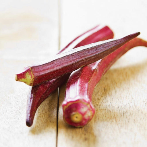 Three fresh, vibrant red okra pods on a light wooden surface.