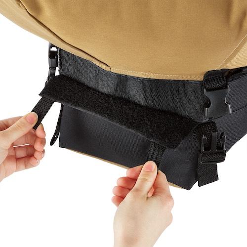Hands fastening a black Velcro strap on a tan and black outdoor gear bag, showing adjustable buckles for secure closure.