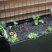 Black raised garden planter with young vegetable plants, colorful pansies, a metal trellis, and wooden background.