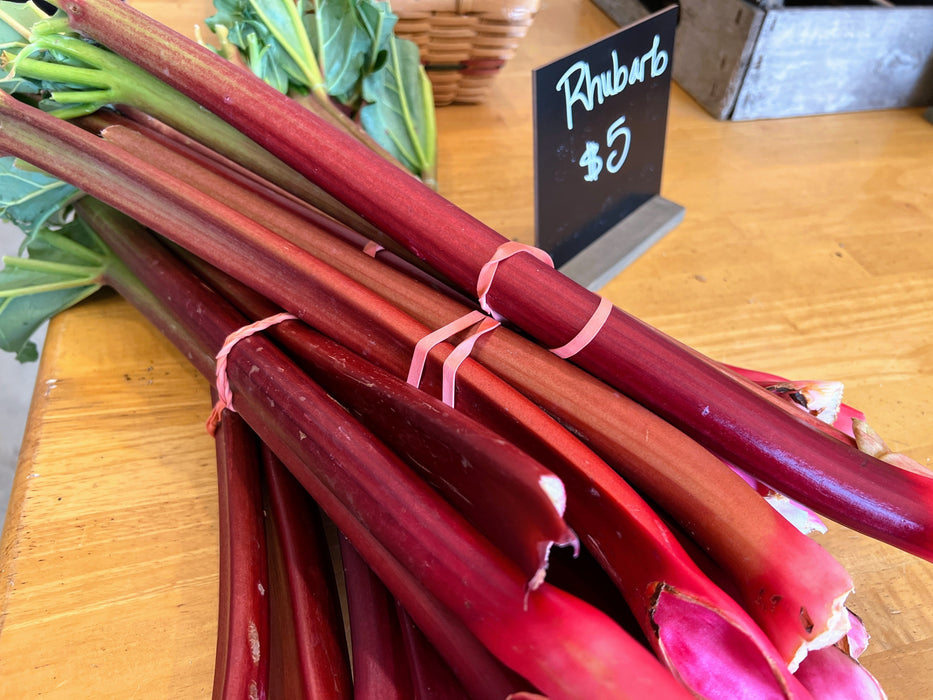 Bundles of fresh red and green rhubarb stalks on a wooden market table with a '$5' price sign.
