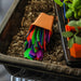 Colorful plant markers spilling from a terracotta pot onto potting soil in a black seed starting tray with young seedlings.