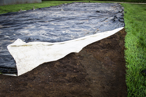 Black weed barrier tarp and white sheet partly covering rich soil, bordered by lush green grass. Landscaping, weed control.