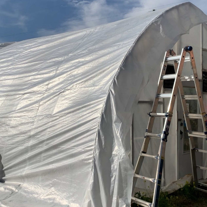 White arched greenhouse or hoop house covered in new plastic sheeting. A-frame ladder for installation. Outdoor farming structure.