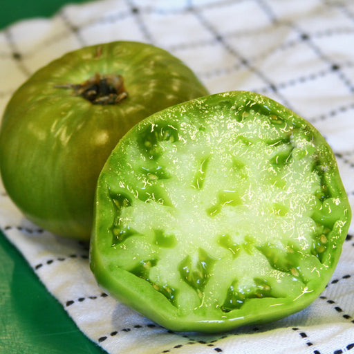 Fresh green tomatoes, one whole and one cut in half, showcasing the vibrant flesh and seeds on a white checkered cloth.