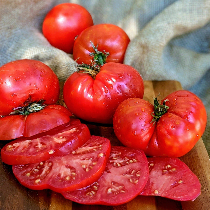 Fresh, ripe red heirloom tomatoes, whole and sliced, glistening with water droplets on a wooden cutting board.