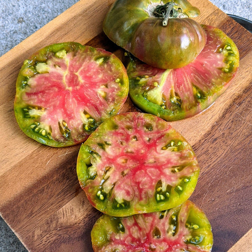 Sliced heirloom tomatoes with green skin and vibrant red flesh on a wooden cutting board. Whole tomatoes nearby.