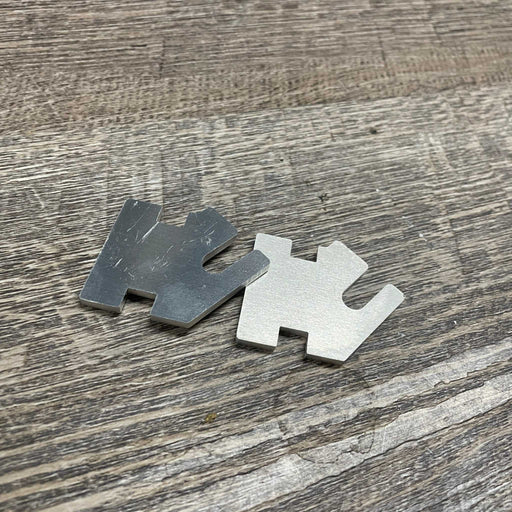Two silver notched metal shims on grey wood grain. Precision spacers for mechanical alignment.
