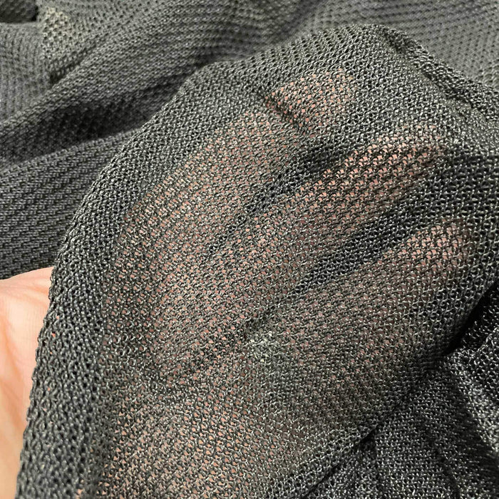 Hand behind sheer dark gray open-knit fabric, showcasing its transparent, breathable mesh texture.