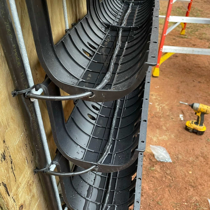 Vertical black plastic cable management trays on wood, holding wet wires and conduit. Construction tools nearby on dirt.