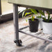 Olive green mobile raised planter with potted plants on a metal grid shelf in a nursery.