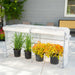 White portable greenhouse protecting green and yellow-orange potted plants on a patio. Ideal outdoor garden plant cover.
