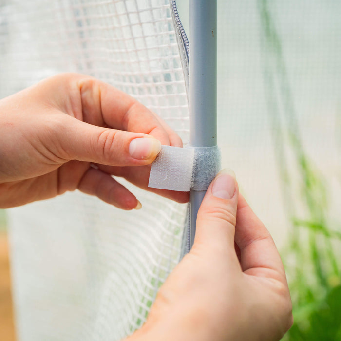 Hands securing a translucent greenhouse cover to a frame pole with a white Velcro strap. Garden setup.