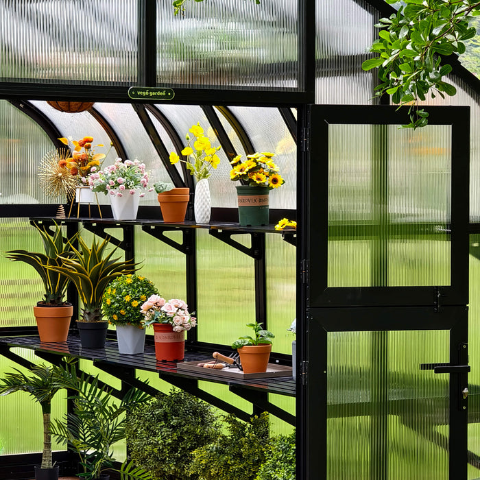 Vego Garden greenhouse with black frame, fluted panels, and shelves displaying potted plants, colorful flowers, and gardening tools.