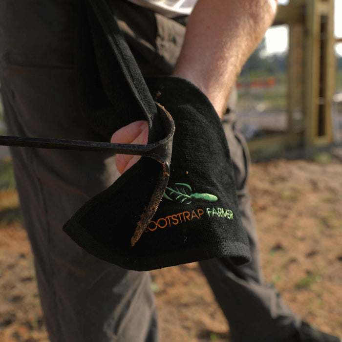 Dirty gardening hoe resting on a black Bootstrap Farmer towel with green logo. Farmer's hand in garden.
