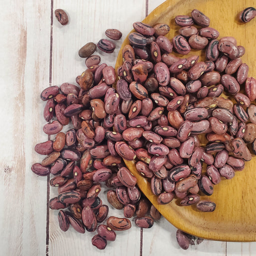 Dried maroon and black striped beans on a rustic wooden plate, spilling onto a light wood table. Healthy legumes, cooking.