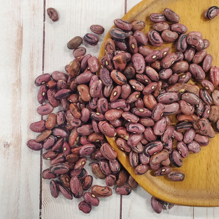 Dried maroon and black striped beans on a rustic wooden plate, spilling onto a light wood table. Healthy legumes, cooking.