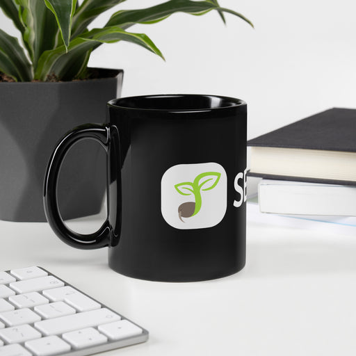 Black coffee mug with green sprouting seed logo on a white desk, near a plant, keyboard, and stacked books.