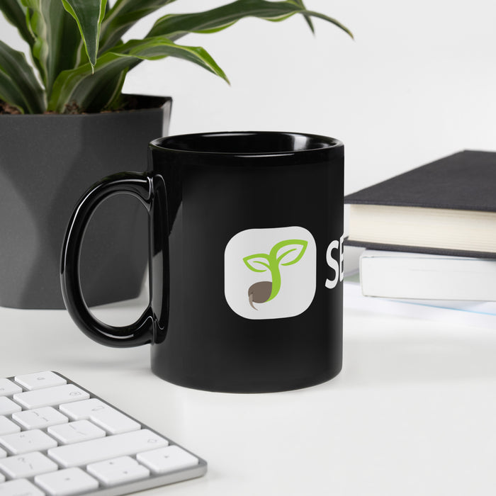 Black coffee mug with green sprouting seed logo on a white desk, near a plant, keyboard, and stacked books.