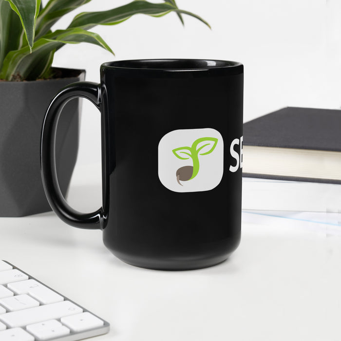 Black coffee mug with green sprout logo, on a desk with a plant, keyboard, and books.
