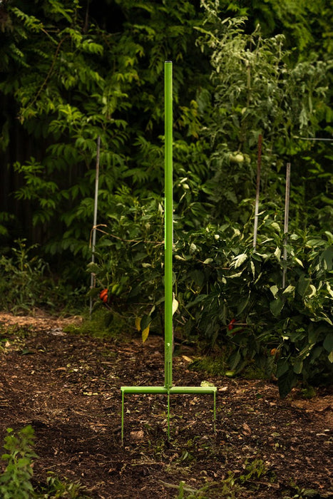 Green broadfork garden tool stuck in rich garden soil, with lush vegetable plants in background.