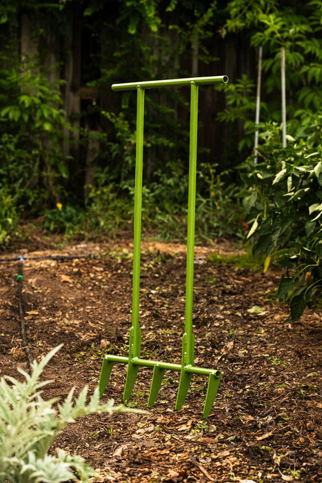 Green broadfork garden tool with four tines, standing upright in mulched garden soil for deep cultivation.