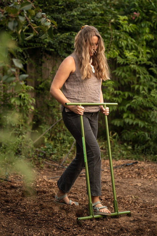Woman using green "Mindful Farmer" garden tool to cultivate soil. Lush outdoor garden background.