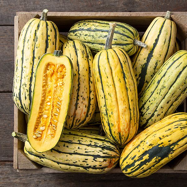 Fresh delicata winter squash in a wooden crate, one cut lengthwise revealing seeds. Yellow and green stripes.