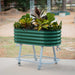 Green corrugated steel mobile raised garden bed on wheels with diverse plants. Features lower shelf and handle. Ideal patio planter.
