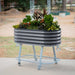 Gray corrugated metal mobile raised garden bed on wheeled light blue stand with lower shelf, full of colorful plants.