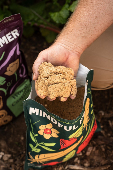 Hand scooping granular organic fertilizer from a Mindful Farm bag for healthy plants. Worm castings bag visible.